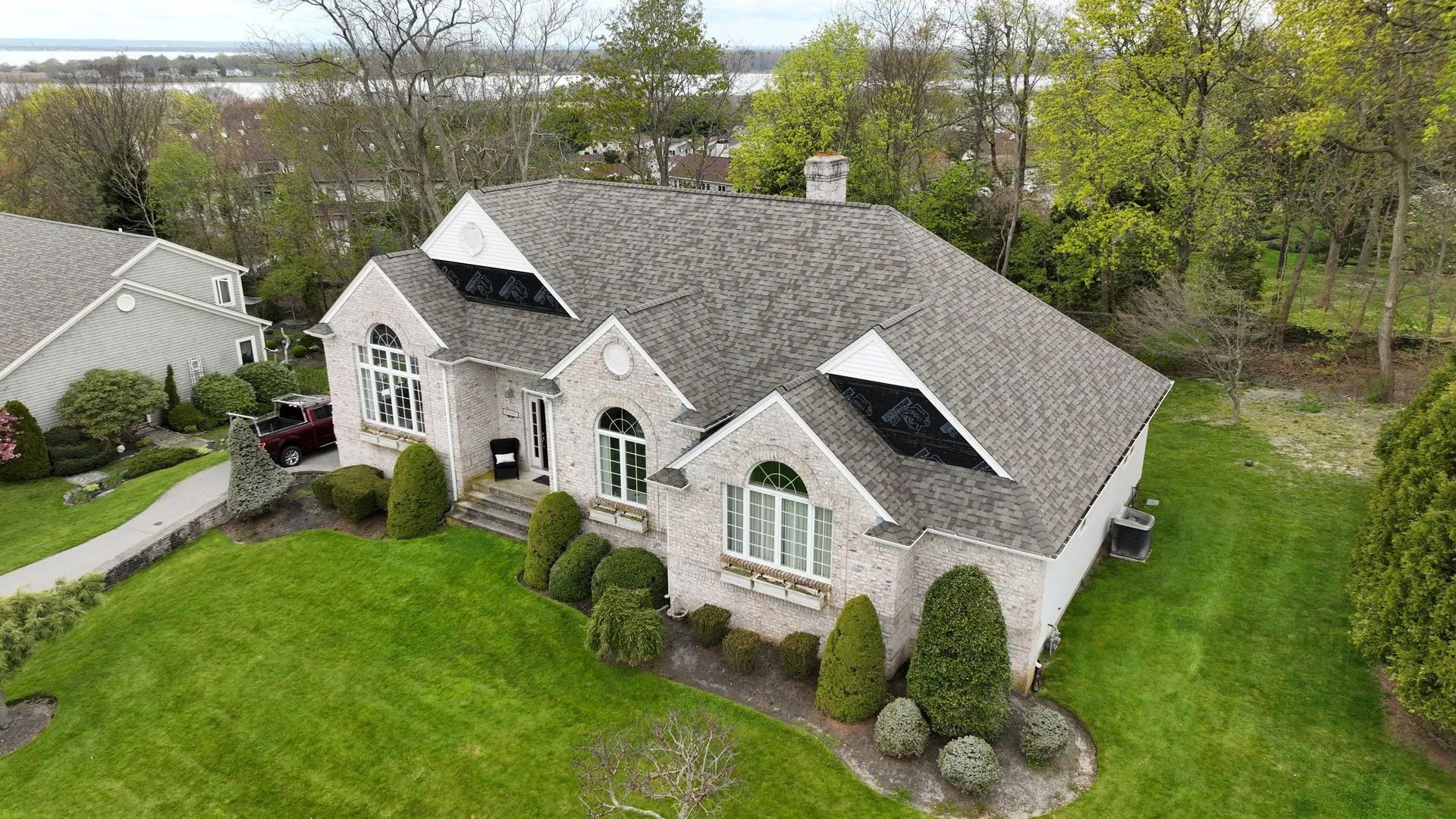 An aerial view of a light-colored brick house with a dark shingled roof, surrounded by a green lawn and trees.