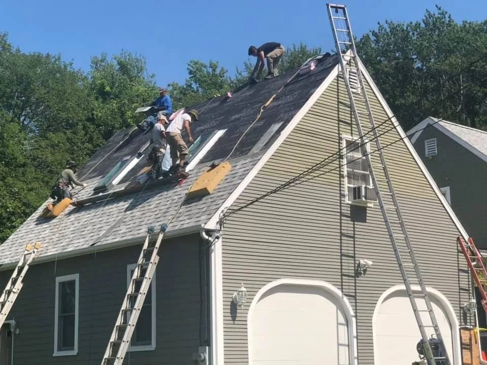 Workers in hard hats perform roofing work on a residential house on a sunny day, using ladders and installing shingles.