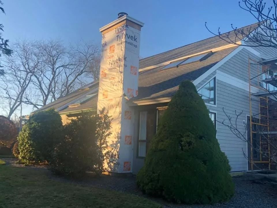 A chimney wrapped in tan, branded protective paper attached to the side of a grey-sided house with scaffolding nearby.