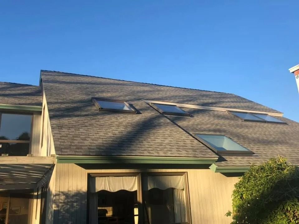A house with a large, grey shingled roof featuring four skylights under a clear blue sky.