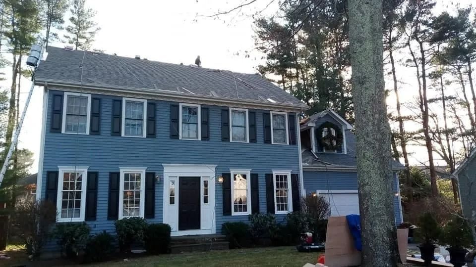 A two-story blue colonial-style house with black shutters, a white front door, and a tree-filled yard at dusk.