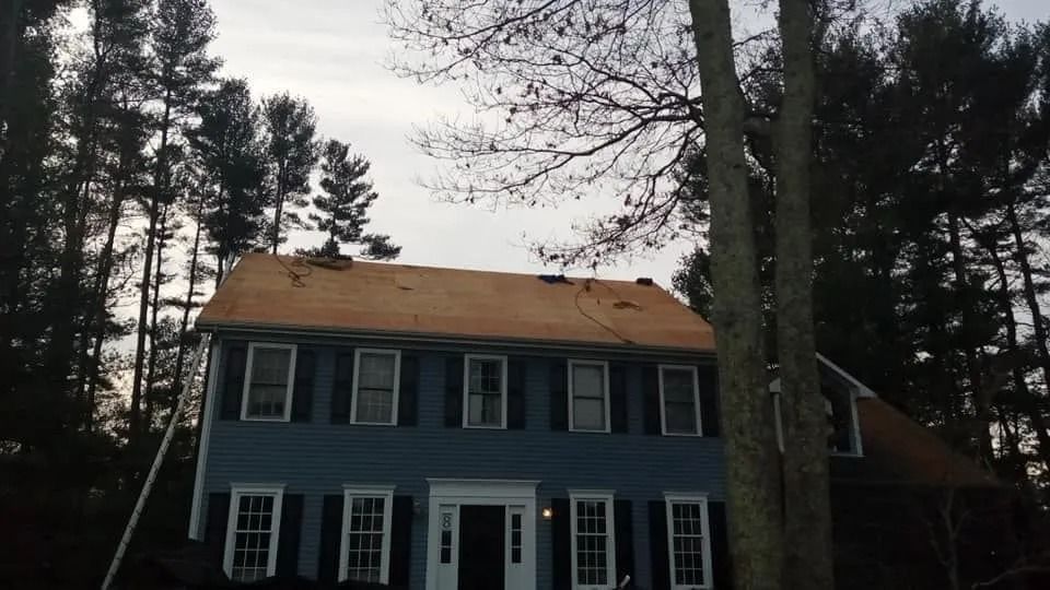 A blue two-story colonial house with shutters and a roof currently undergoing replacement, surrounded by tall pine trees.