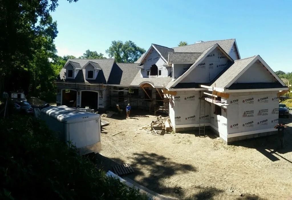 A large house under construction with gray sheathing and a shingled roof, set on a dirt lot with nearby trees.