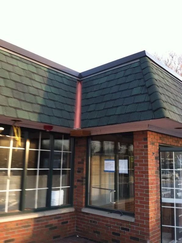 Exterior view of a brick building corner with green shingled roofing and a central copper-colored downspout.