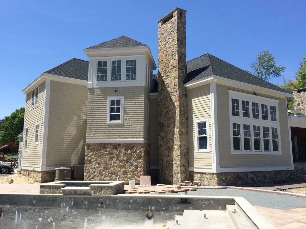 A beige house with a stone chimney and foundation under a clear blue sky, featuring a pool under construction in front.
