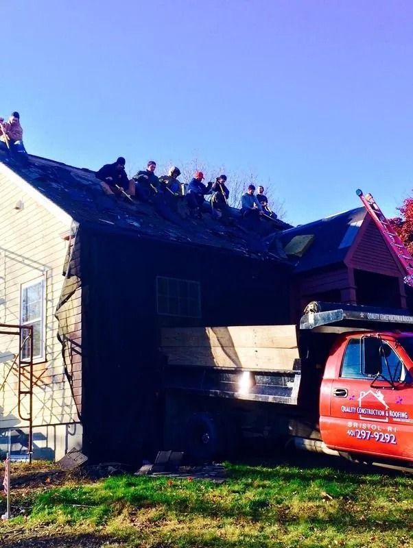 A team of workers stands on a residential roof removing shingles, with a red company truck parked alongside the house.