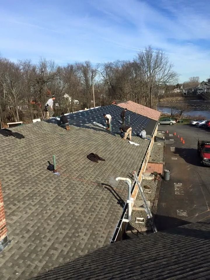 Construction crew repairing a shingled roof on a sunny day near a body of water and parking lot.