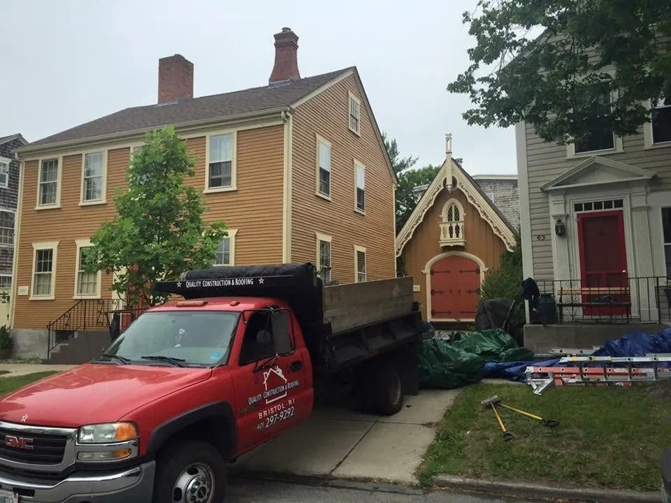 A red GMC dump truck parked on a residential street between a yellow house and a small carriage house with a red door.