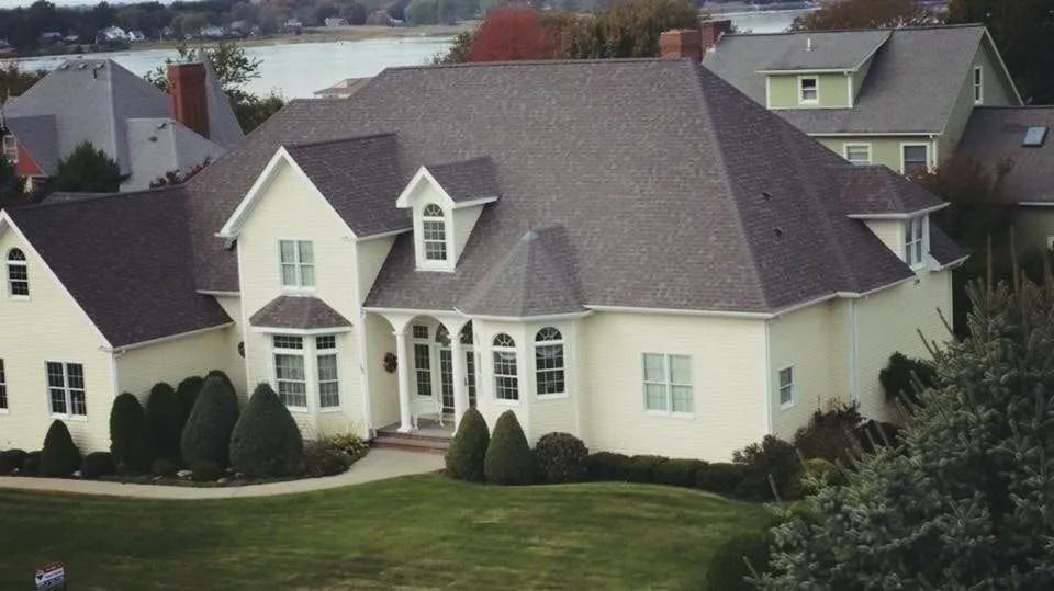 An aerial view of a large, cream-colored house with a dark gray shingled roof, set in a grassy yard near a body of water.