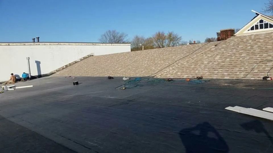 Construction workers install light-colored tiles on a large, sloped roof section adjacent to a flat, dark roof surface.