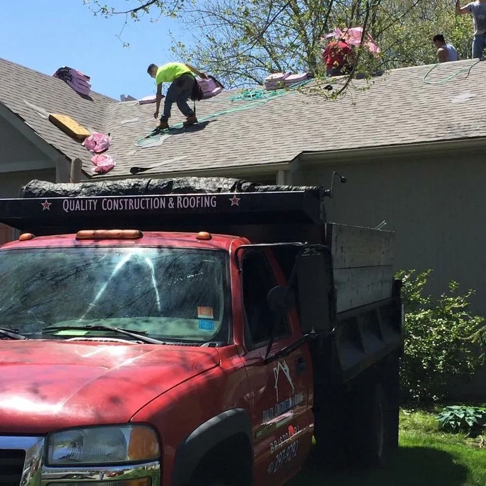 Workers replace shingles on a residential roof next to a red Quality Construction & Roofing dump truck.