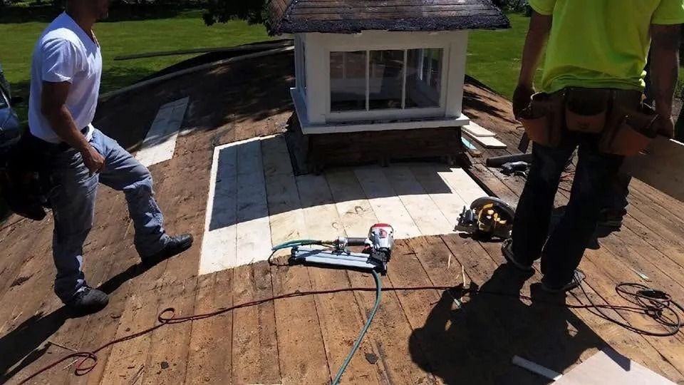Two construction workers installing new plywood roof decking around a small rooftop cupola on a sunny day.