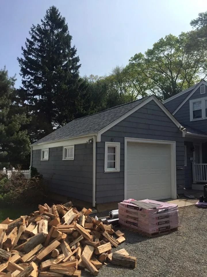 A blue-sided garage with a white door, a stack of firewood in the foreground, and a large pine tree in the background.