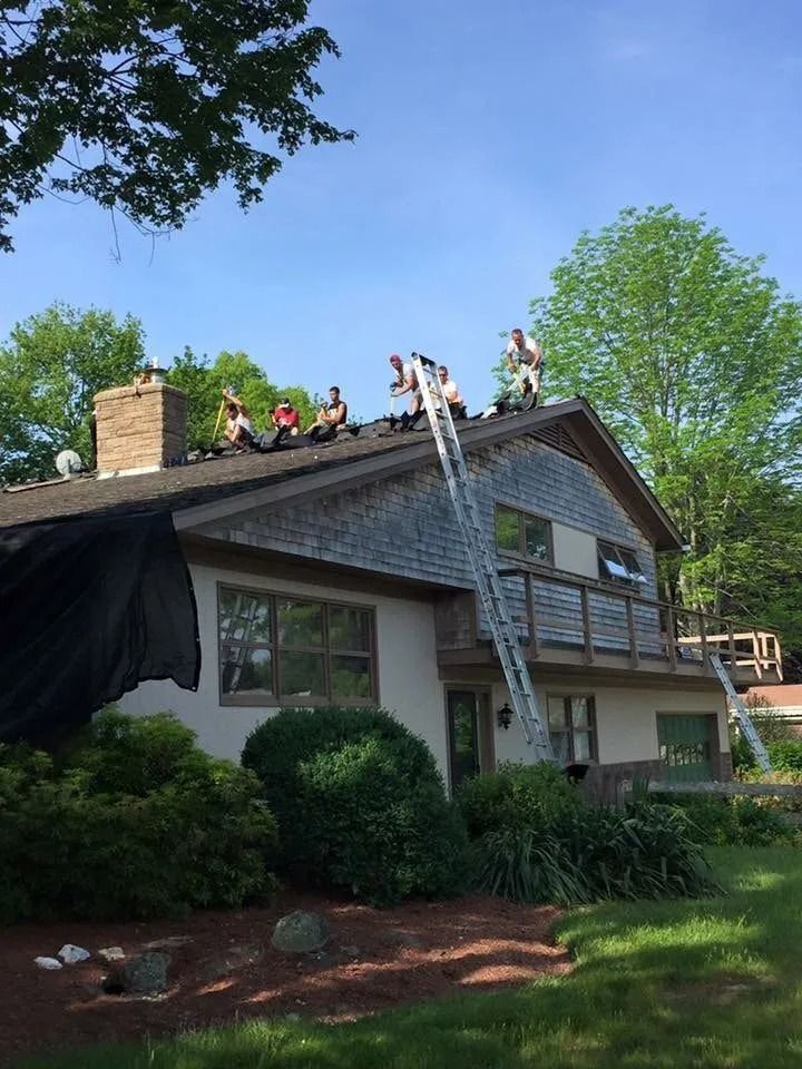 A crew of roofers work on the shingled roof of a two-story residential house on a sunny day.