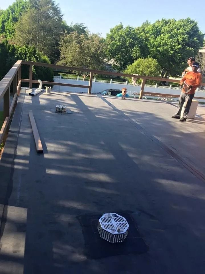 Two people stand on a flat, dark gray roof with a wooden railing, featuring a central circular drainage cover.
