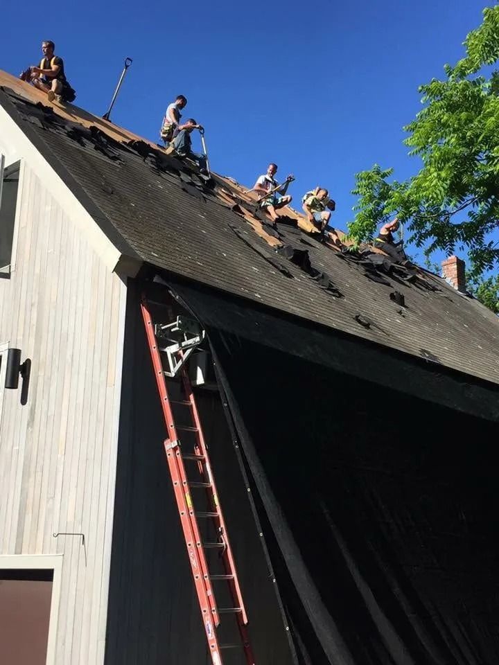 Several people working on a steep residential roof to remove old shingles, with a red ladder leaned against the side.