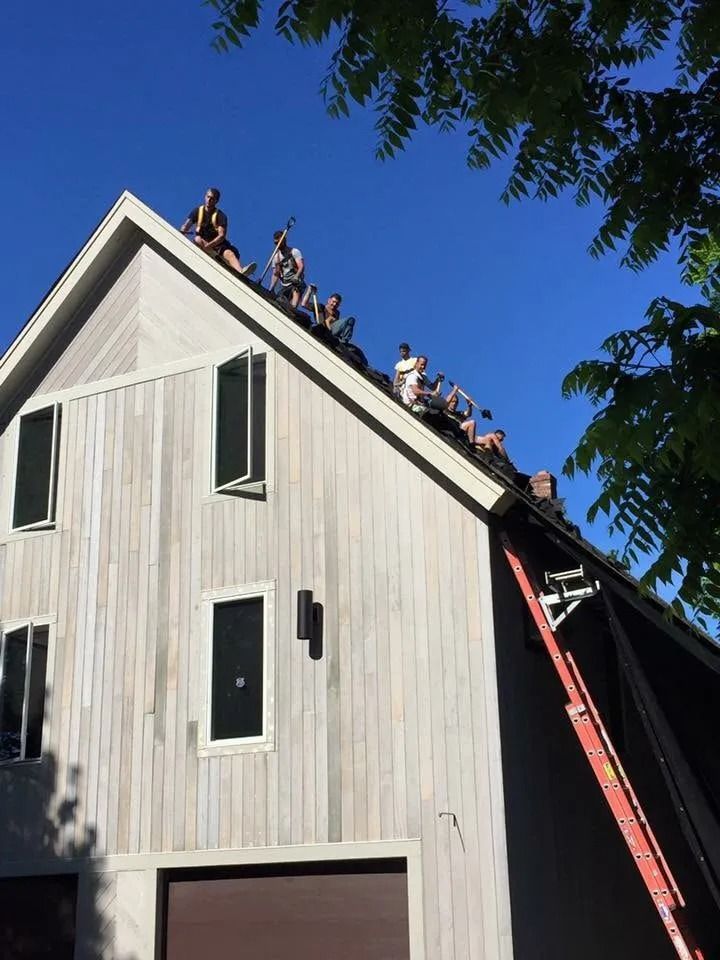 A group of people sit together on the steep roof of a light-colored wooden building under a clear blue sky.
