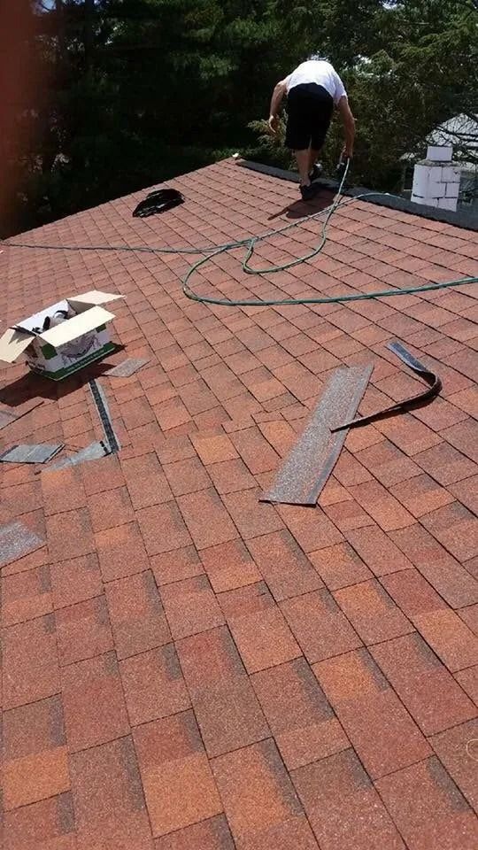 A person repairs a shingled roof with tools, materials, and a hose nearby on a sunny day.