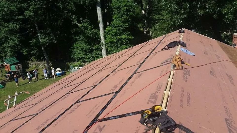 A person’s view from a sloped roof covered in pink ZIP System sheathing with taped seams and a ridge vent being installed.