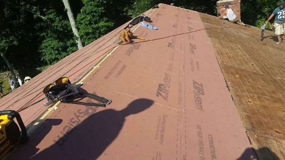 Workers install red ZIP System roofing underlayment on a wooden roof deck next to a brick chimney.