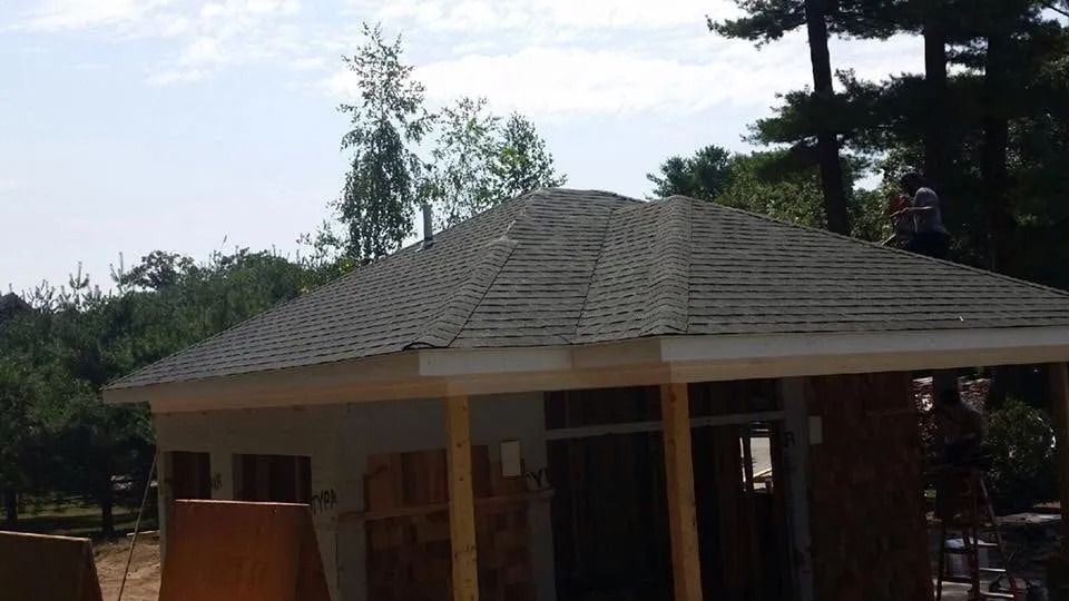 An unfinished structure with a newly shingled roof stands in a wooded area under a partly cloudy sky.