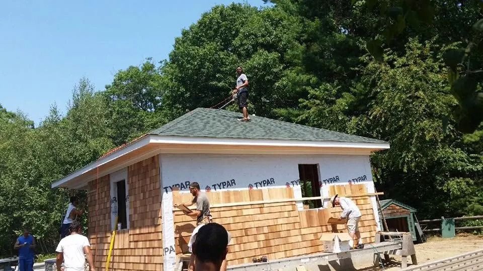 Construction workers apply wood shingle siding to the exterior walls of a small building under a clear blue sky.
