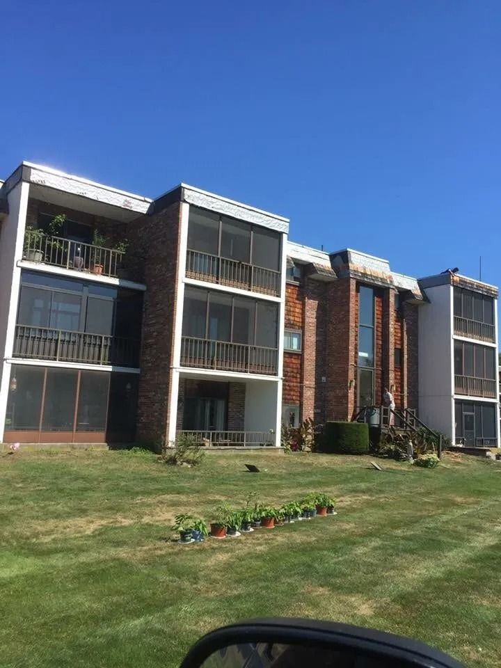 A three-story apartment building with brick siding and screened balconies, surrounded by lawn with small potted plants.