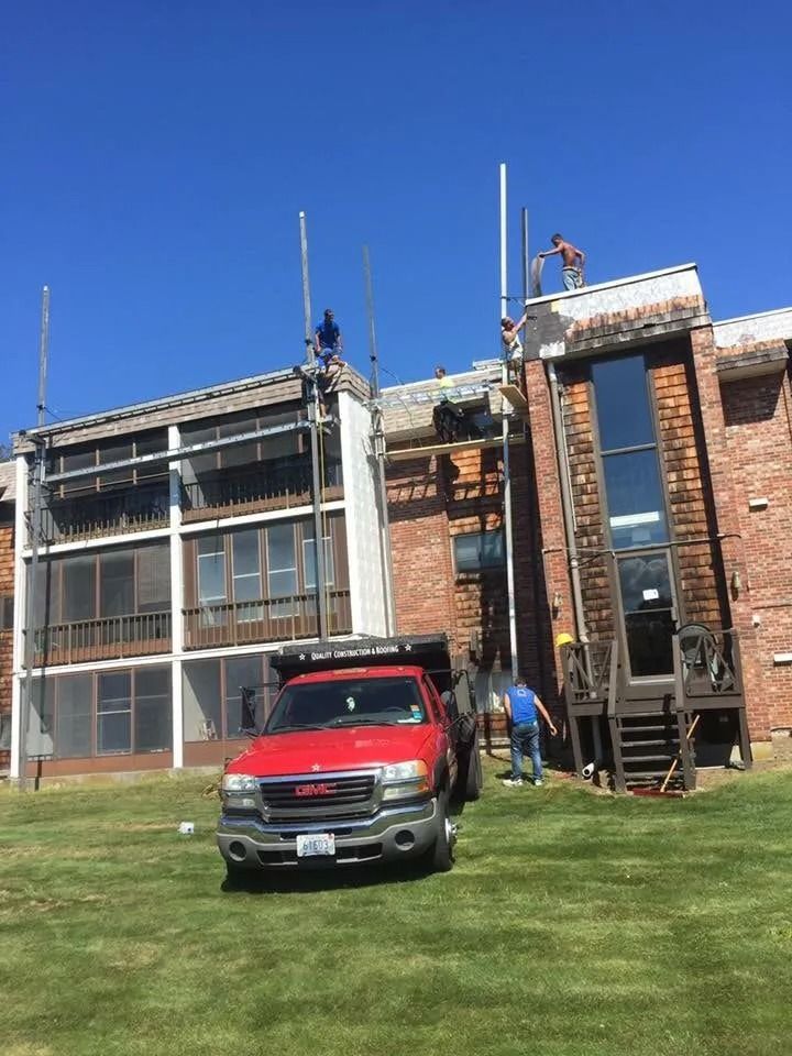 Construction workers on scaffolding repair the roof of a multi-story brick apartment building with a red truck parked nearby.