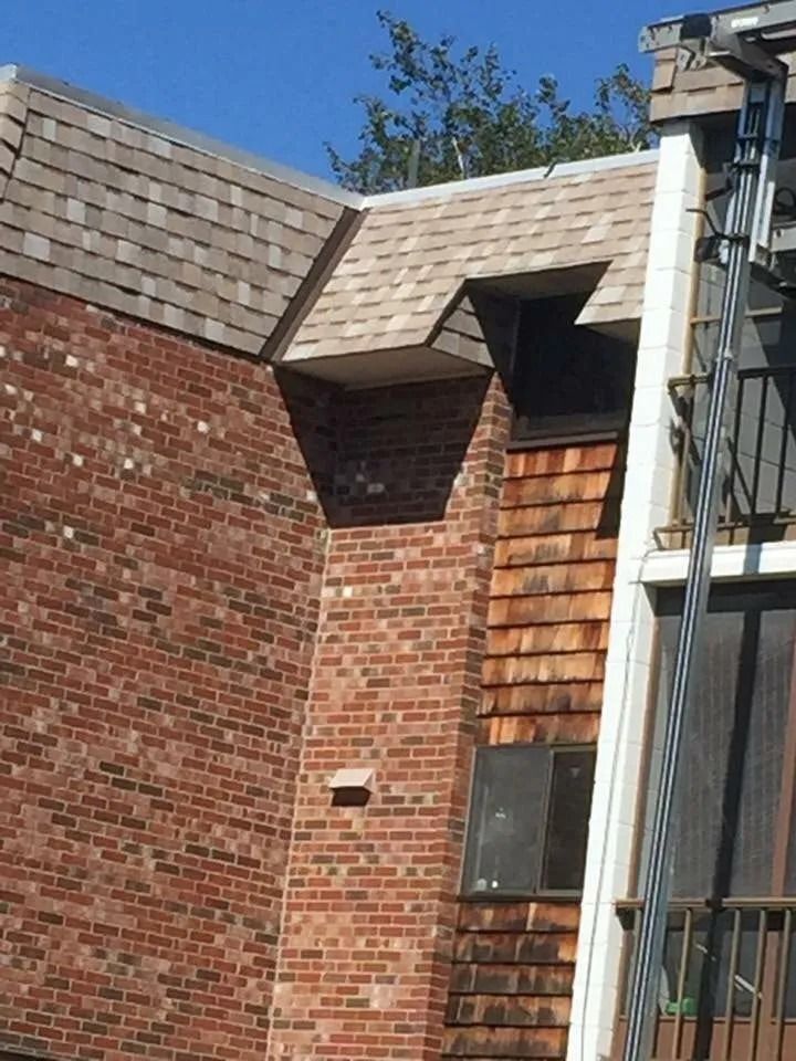 A brick building exterior featuring a complex roofline, cedar shake siding, a window, and a metal ladder on the right.