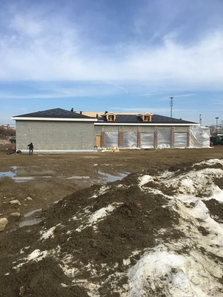 A one-story building under construction with a gray masonry side, a shingled roof, and plastic-covered door openings.
