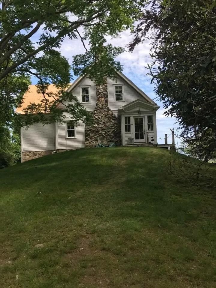 A white house with a stone chimney sits atop a grassy hill, framed by trees under a partly cloudy sky.