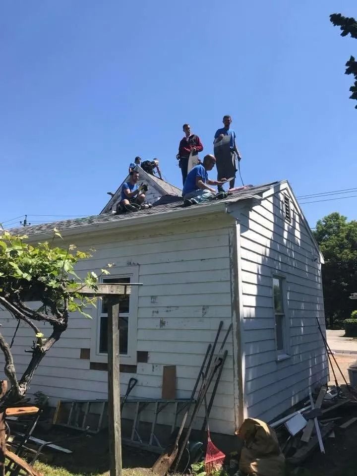 A group of people working on the roof of a small white house on a sunny day.
