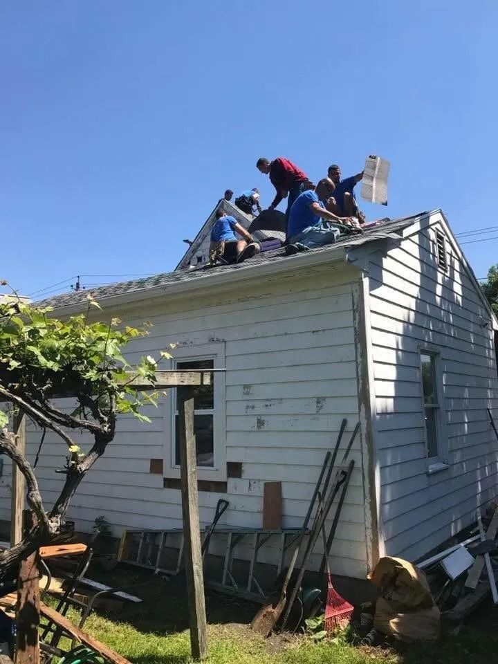 Several people work on the roof of a small white house with wood siding under a clear blue sky.