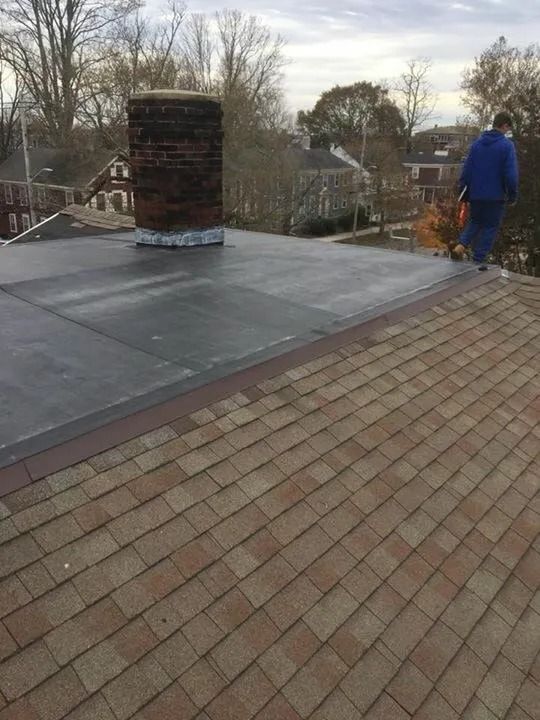 A person in a blue jacket walks on a shingled roof next to a flat, dark roof section and a brick chimney.