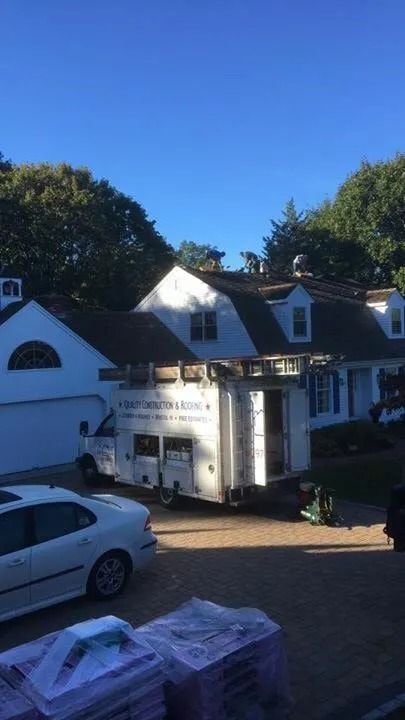 Roofers work on the roof of a white house with a service truck and materials in the driveway.