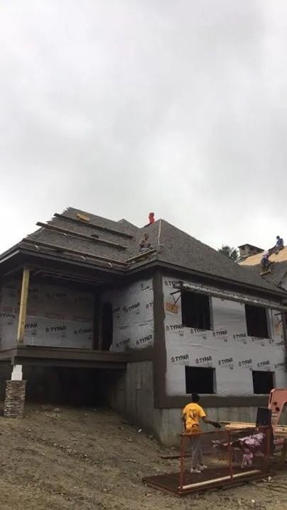 A multi-story house under construction with roof shingles being installed, viewed from a dirt lot on a cloudy day.