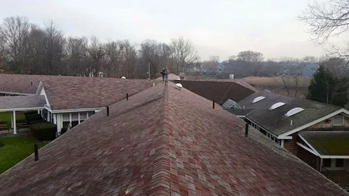 A high-angle view of a brown shingled roof with several vents, stretching toward trees and buildings under a cloudy sky.