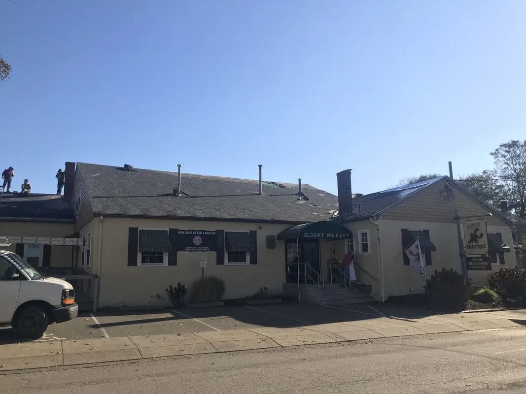 A light-colored, single-story building with a dark shingled roof and roofers working on the left side under a clear sky.
