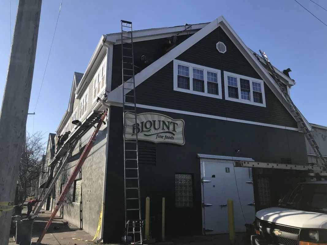 Workers on ladders perform exterior maintenance on a dark-colored building labeled 