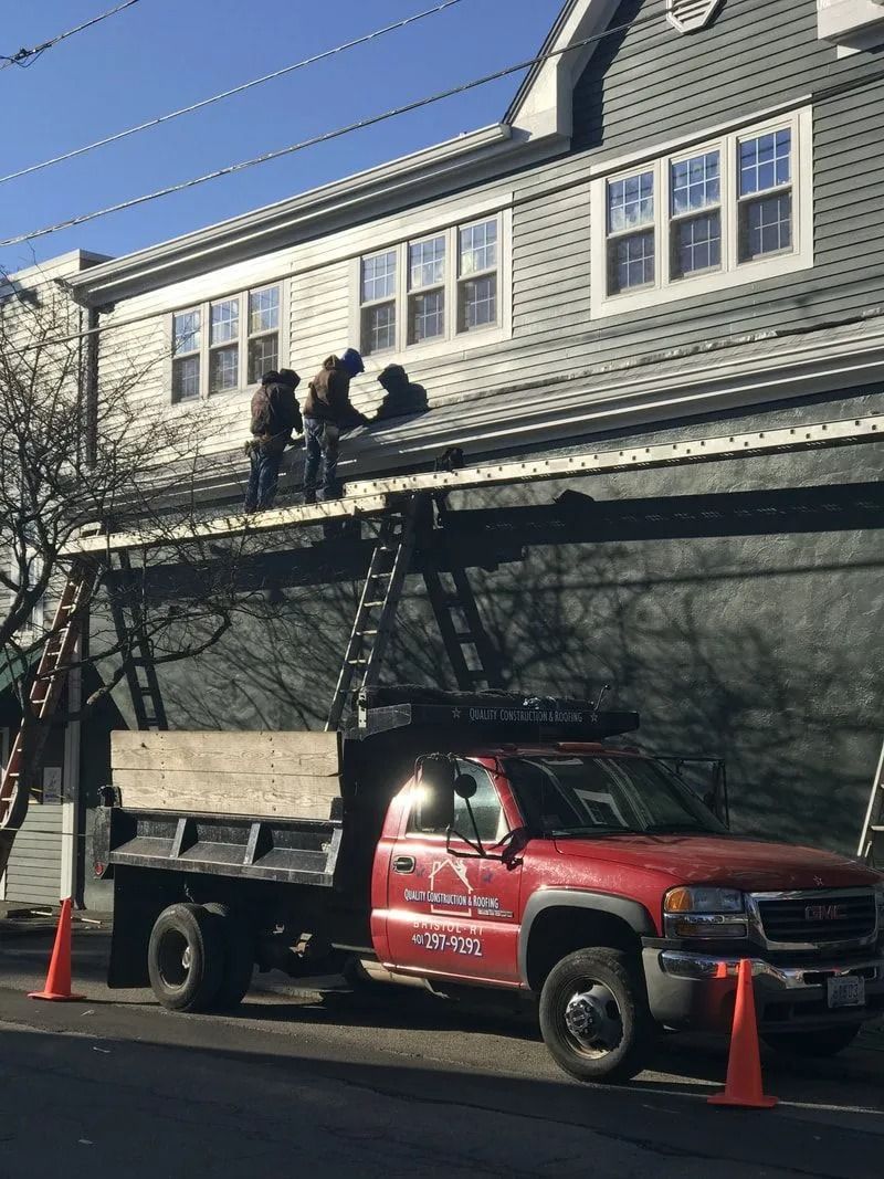 Workers on a ladder repair the roof of a gray house next to a red dump truck with orange cones on a street.
