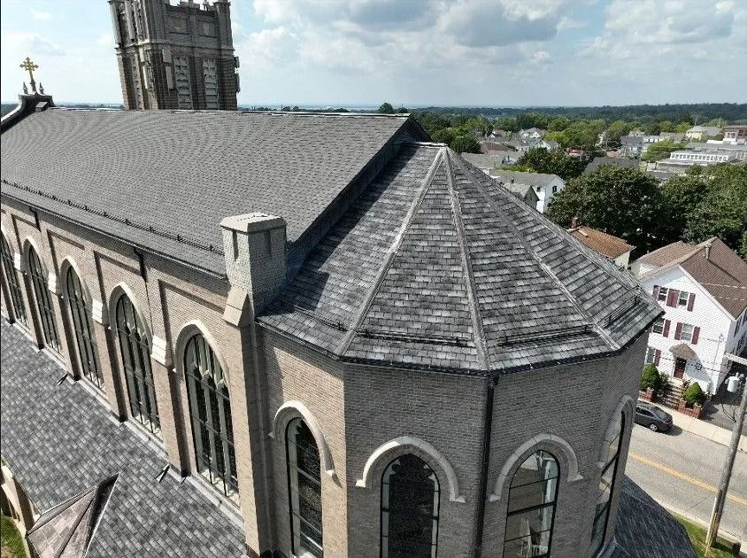 An elevated view of a brick church with a dark gray shingled roof and stained-glass windows overlooking a small town.