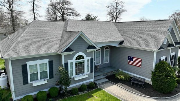 A grey suburban house with a shingled roof, a paved walkway, landscaping, and an American flag displayed on the wall.