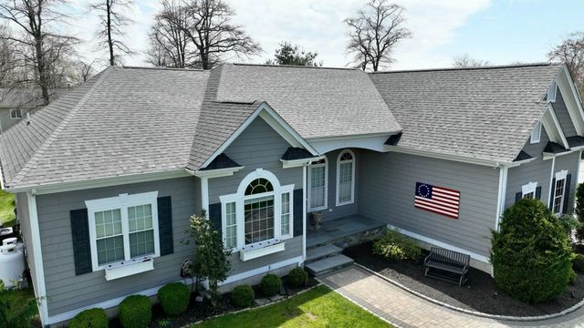 A grey suburban house with a shingled roof, a paved walkway, landscaping, and an American flag displayed on the wall.