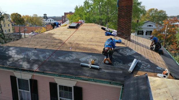 Two workers in blue shirts install black underlayment on the wooden plywood roof of a pink house.