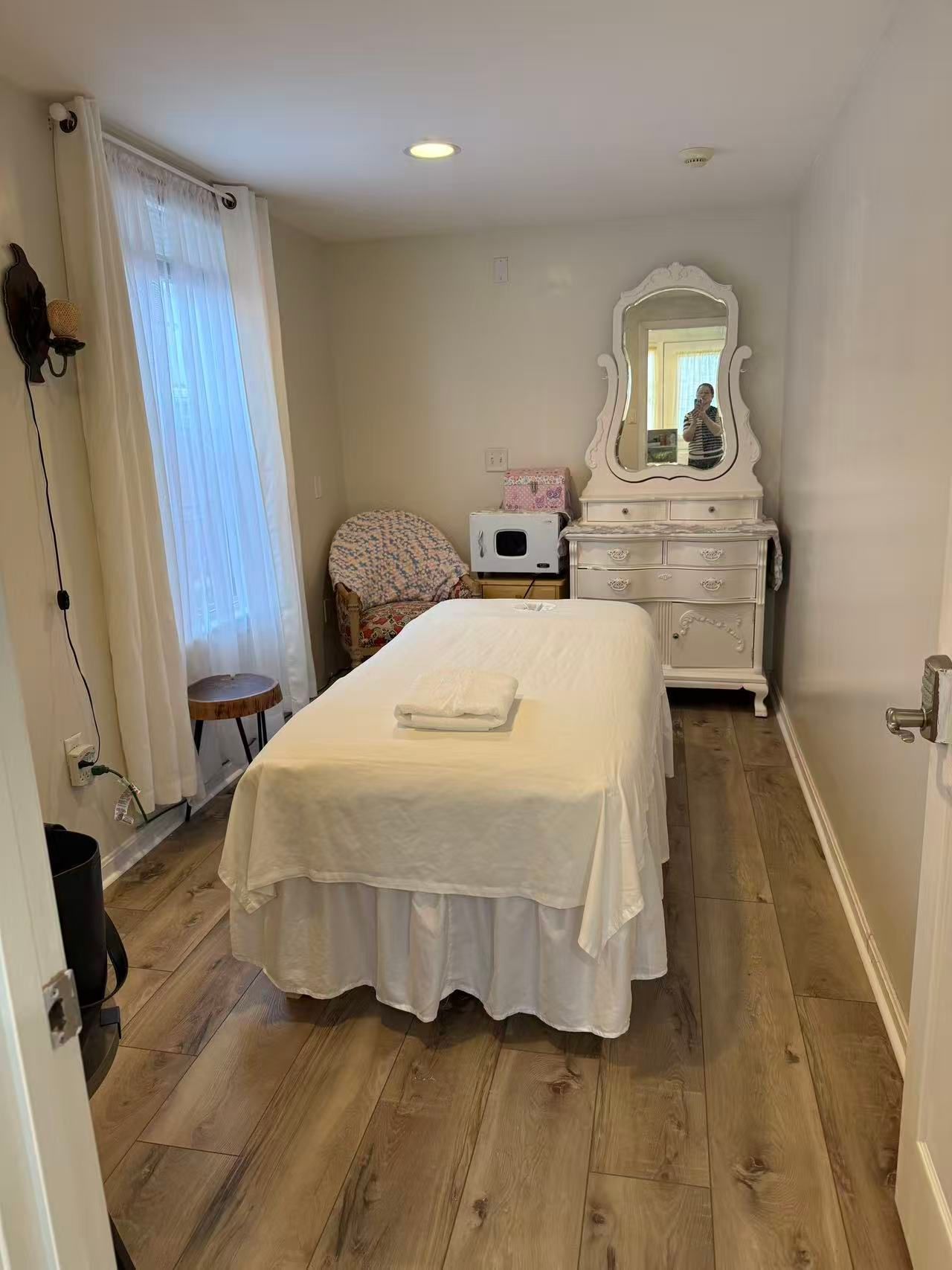 Massage room with bed, chair, dresser, and window. Beige walls and wood-look flooring.