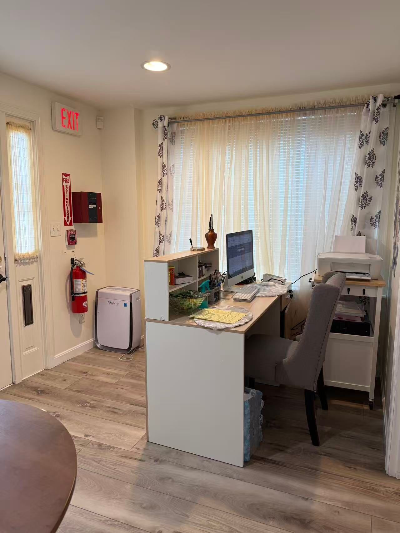 Reception area with white desk, computer, printer, and fire safety equipment near an exit door.
