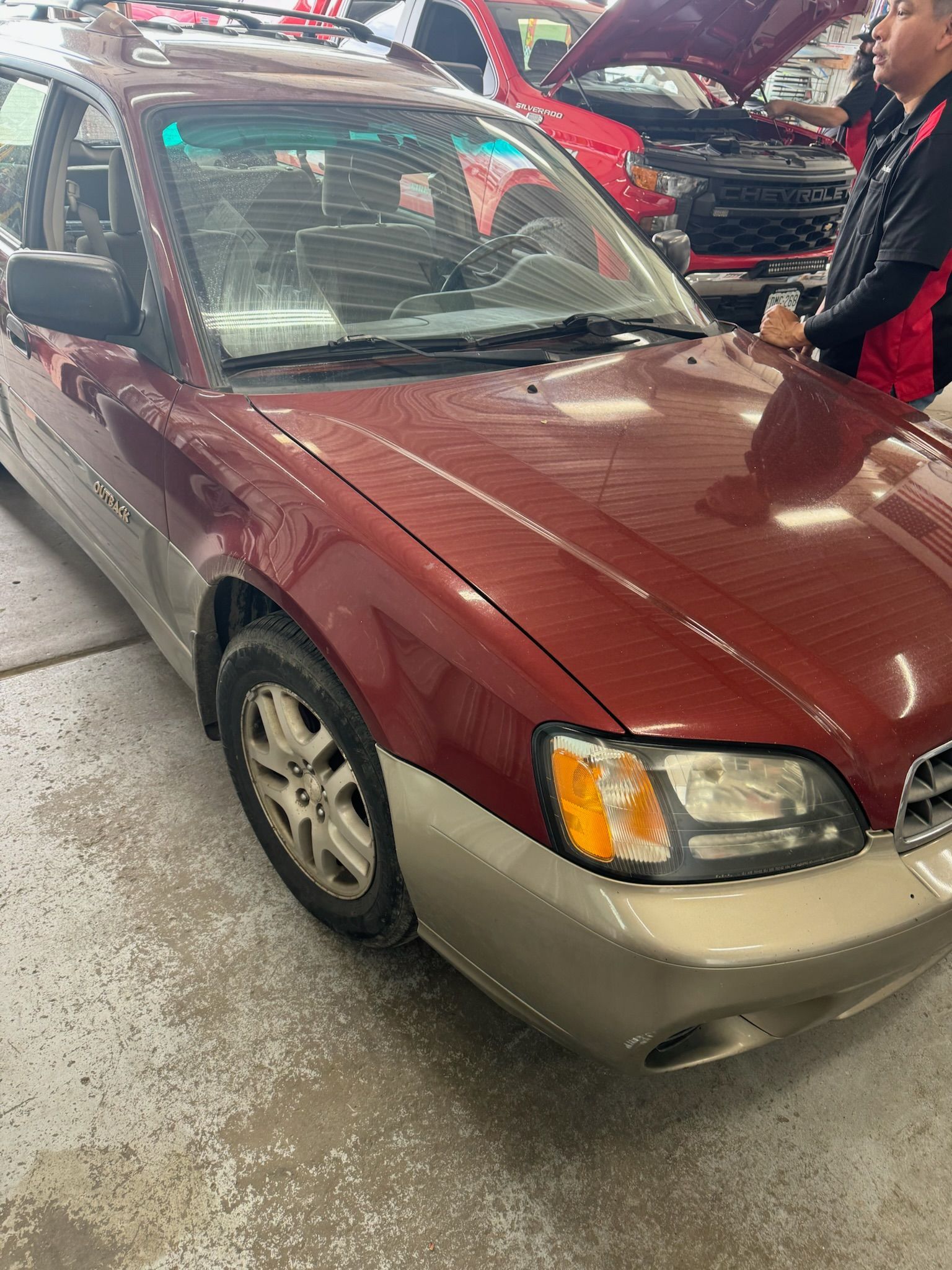 A man is standing next to a red car with the hood up in a garage.