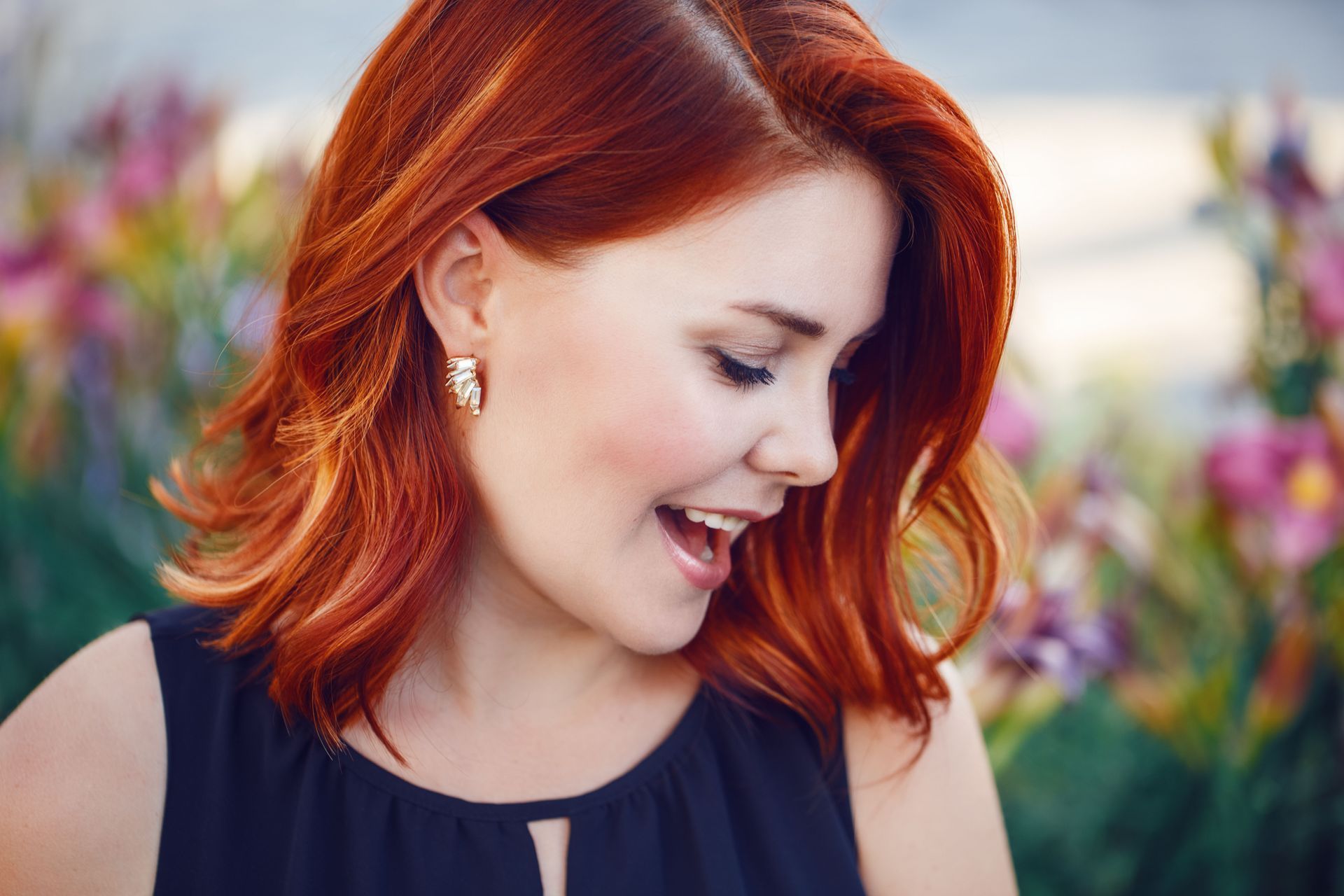 Woman with red hair smiling, looking down; wearing a black dress, flowers in background.