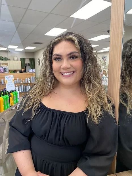 Woman with curly hair smiles, stands in a salon with hair products in the background.