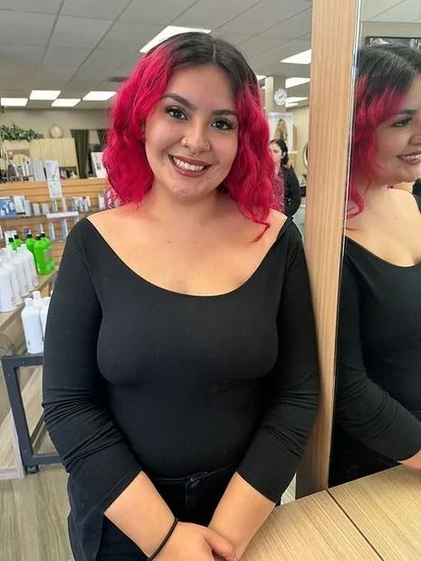 Woman with pink hair smiles, leaning on a mirror in a salon.